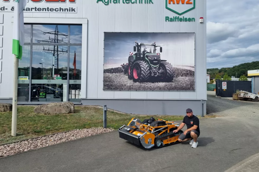Mitarbeiter präsentiert eine ferngesteuerte Mähraupe vor dem Gelände eines Landtechnik-Händlers.
